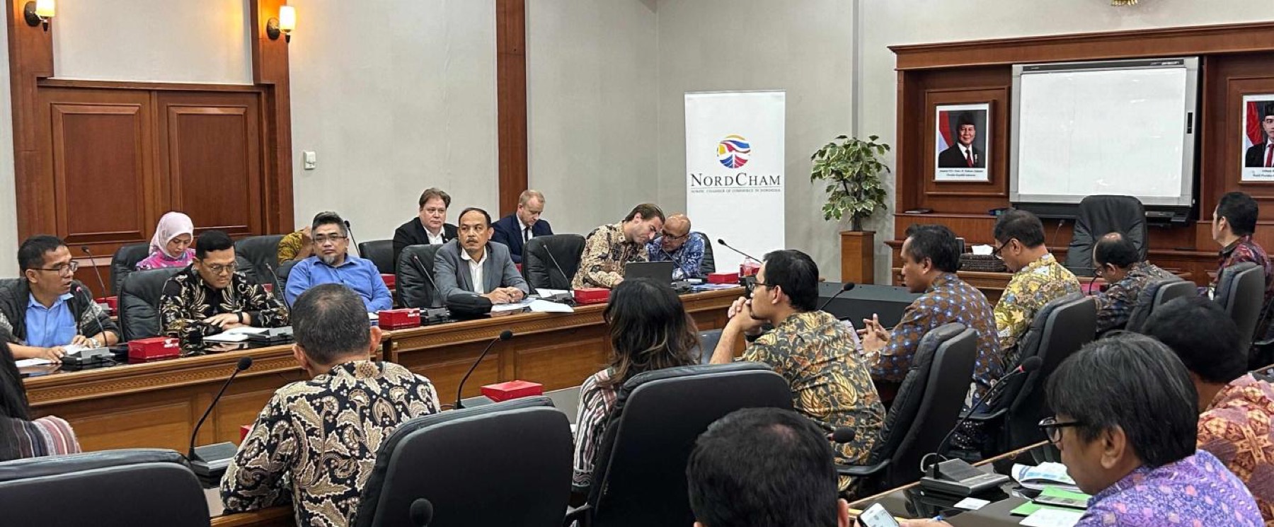 A meeting in a panelled government committee room, a NordCham banner at the head of the table